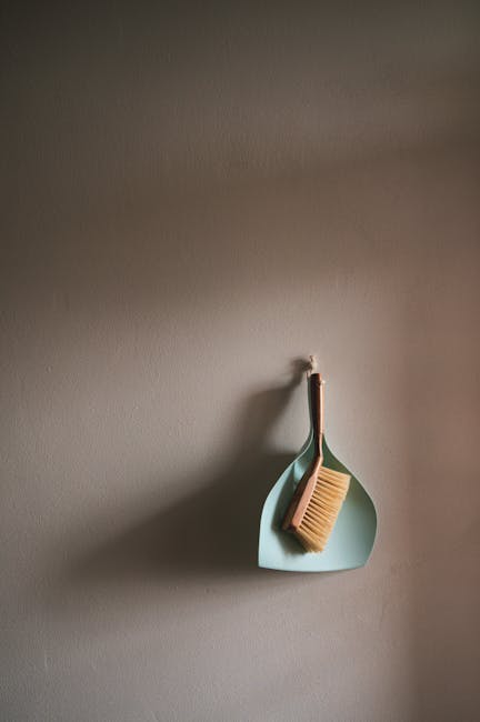 A close-up of a pale blue silicone wall hook attached to a smooth, neutral-toned painted wall in a domestic setting. Hanging from the hook is a small, flat, dustpan-shaped container with a natural woo