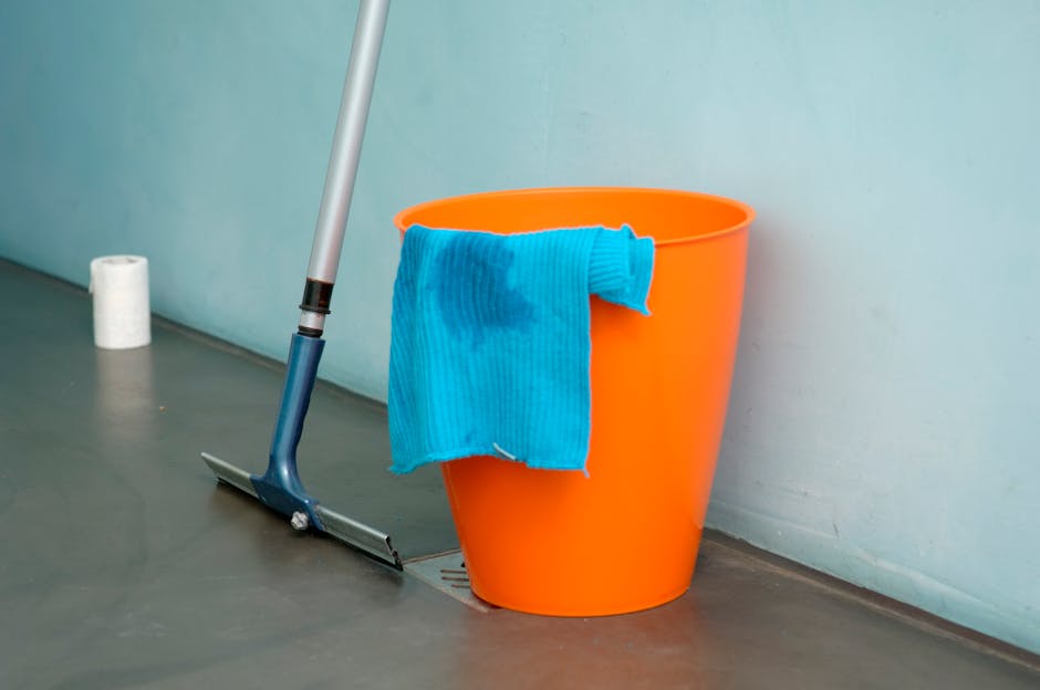 A close-up of a pale blue silicone wall hook attached to a smooth, neutral-toned painted wall in a domestic setting. Hanging from the hook is a small, flat, dustpan-shaped container with a natural woo