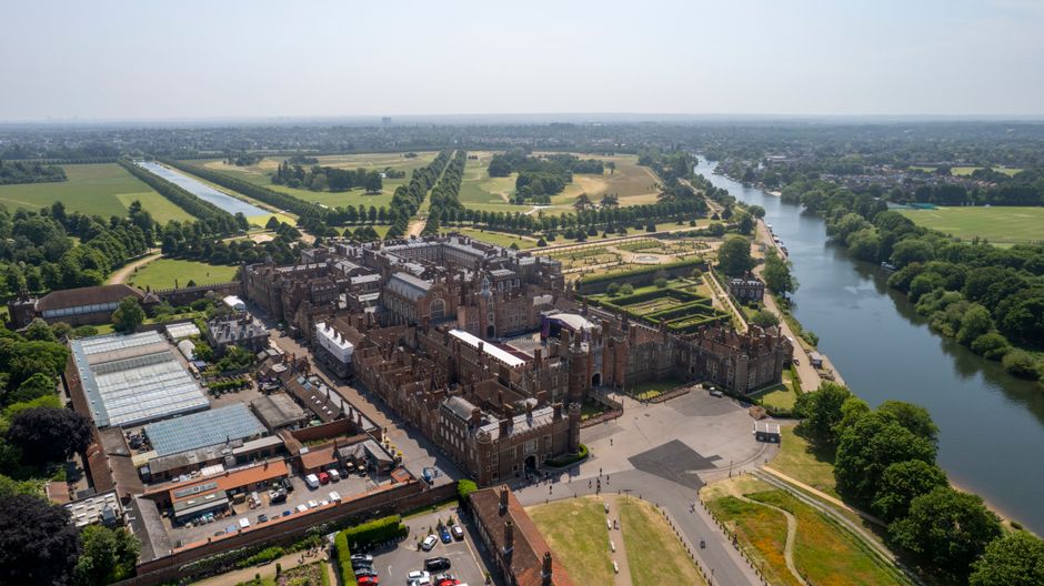 Aerial view of Hampton Court Palace surrounded by well-maintained formal gardens, symmetrical hedges, and green lawns, with a river running along one side. The grand historical building features detai